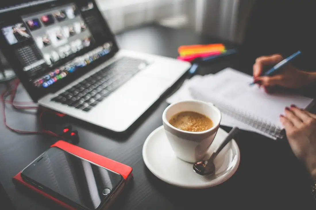 A hand taking notes beside a laptop, espresso and phone while comparing Thai course formats