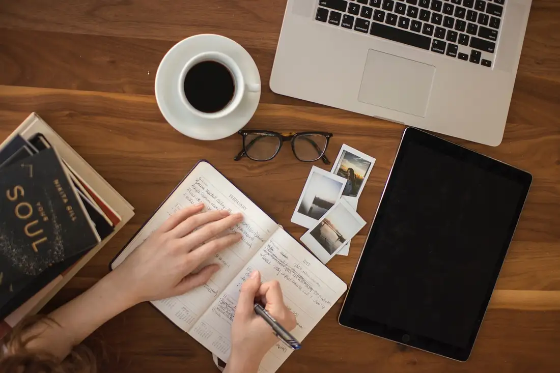 Overhead view of hands writing in a planner beside a laptop, coffee and glasses