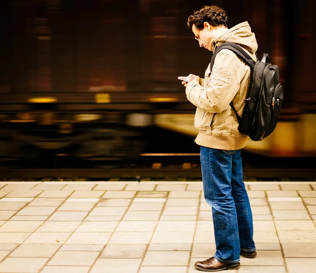 A man with a backpack reading his phone as a train blurs past the platform