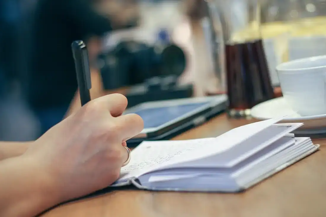 A hand writing in a small notebook beside a coffee, planning how to learn Thai